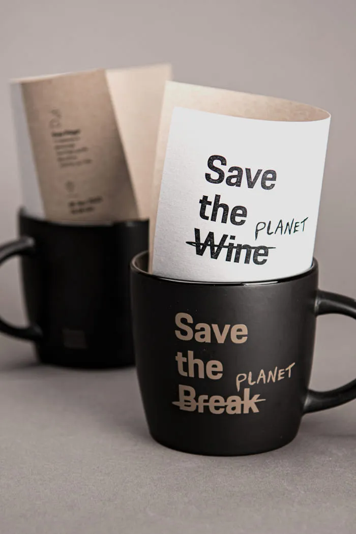 Zwei schwarze Tassen mit goldener Aufschrift stehen auf einem grauen Hintergrund. In der vorderen Tasse steckt ein gefaltetes Blatt mit dem Schriftzug Save the Wine – Wine durchgestrichen und ersetzt durch Planet, auf der Tasse selbst steht Save the Break – wiederum ersetzt durch Planet.
